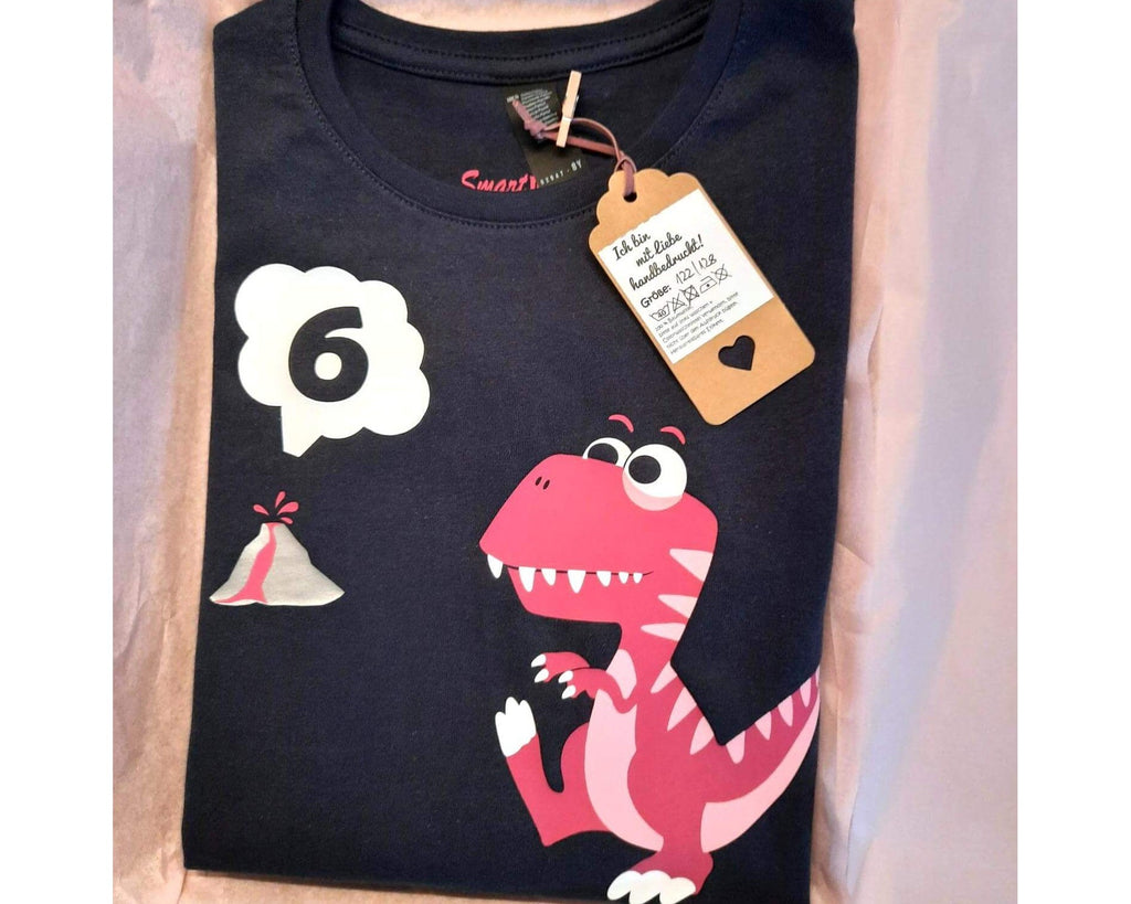 navi farbenes Geburtstagsshirt in Verpackung mit Etikett, bedruckt mit T Rex in rosa-pink, mit Name und Zahl