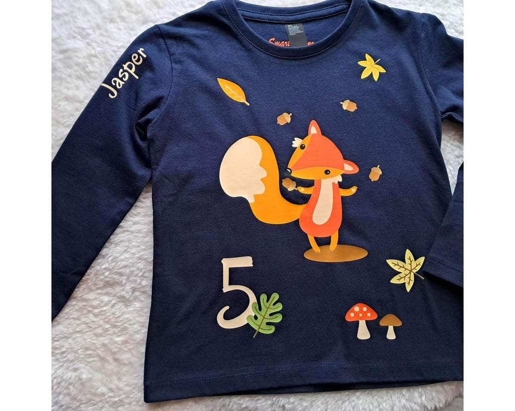 navi farbenes Geburtstagsshirt für Kinder, bedruckt mit süßem Fuchs, der Eicheln jongliert und herbstlicher Szene, Name und Zahl, als Flatlay