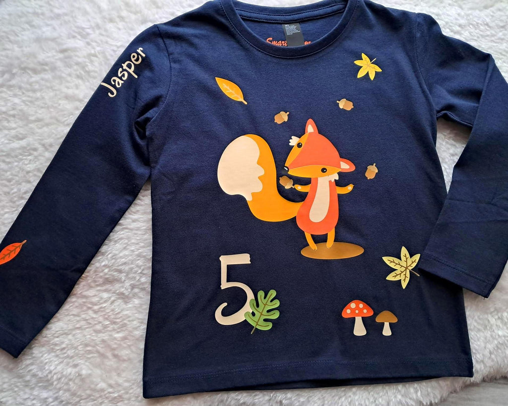 navi farbenes Geburtstagsshirt für Kinder, bedruckt mit süßem Fuchs, der Eicheln jongliert und herbstlicher Szene, Name und Zahl, als Flatlay