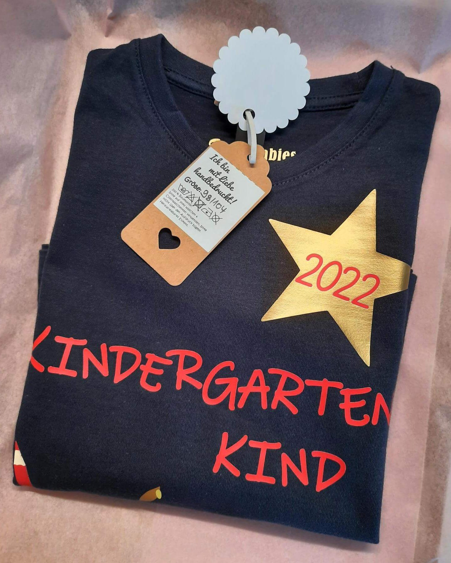 Verpackung mit Etikett navi Kindergartenshirt mit 
Name und Jahreszahl, roter Aufdruck mit schimmerndem gold